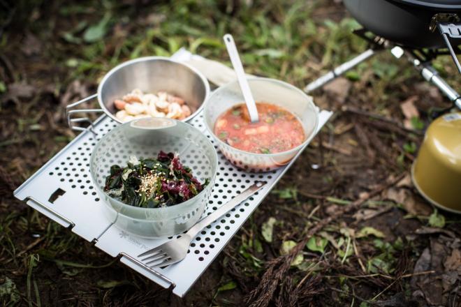 An outdoor meal of salad, pasta, and tomato soup in metal bowls on a perforated metal tray.