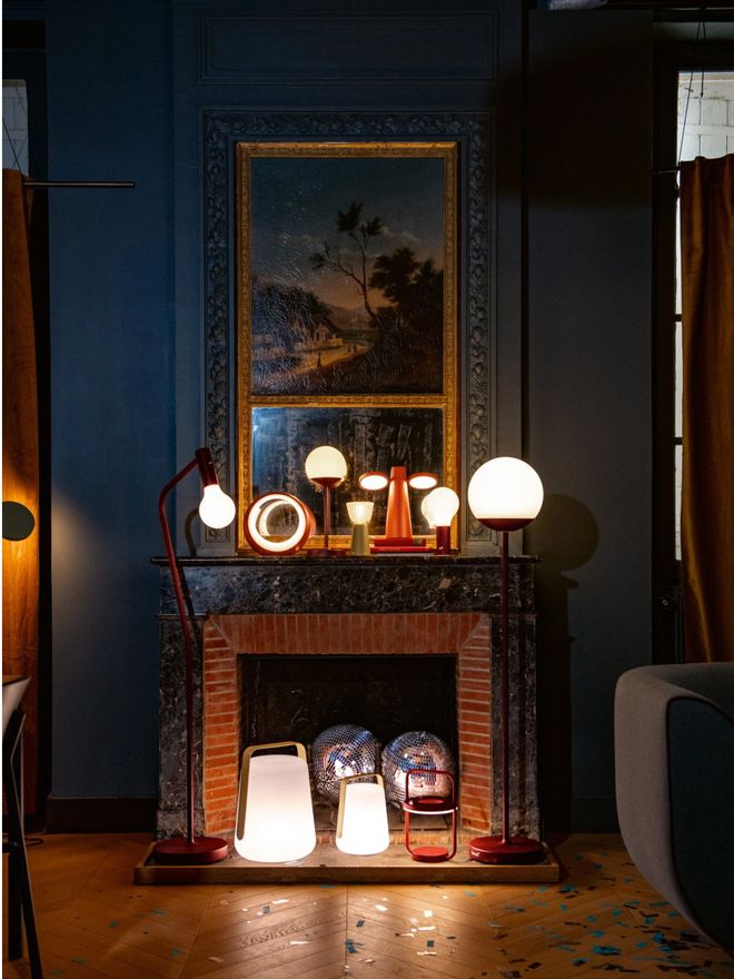 Fermob portable lamps on a fireplace mantel and the floor in a dark blue room with a gilt-framed painting on the wall.
