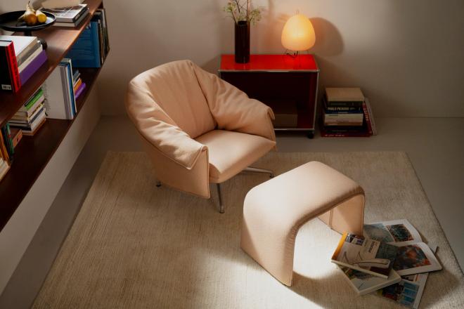 A blush-toned lounge chair and matching ottoman on a rug, with a low bookshelf and a red side table behind.