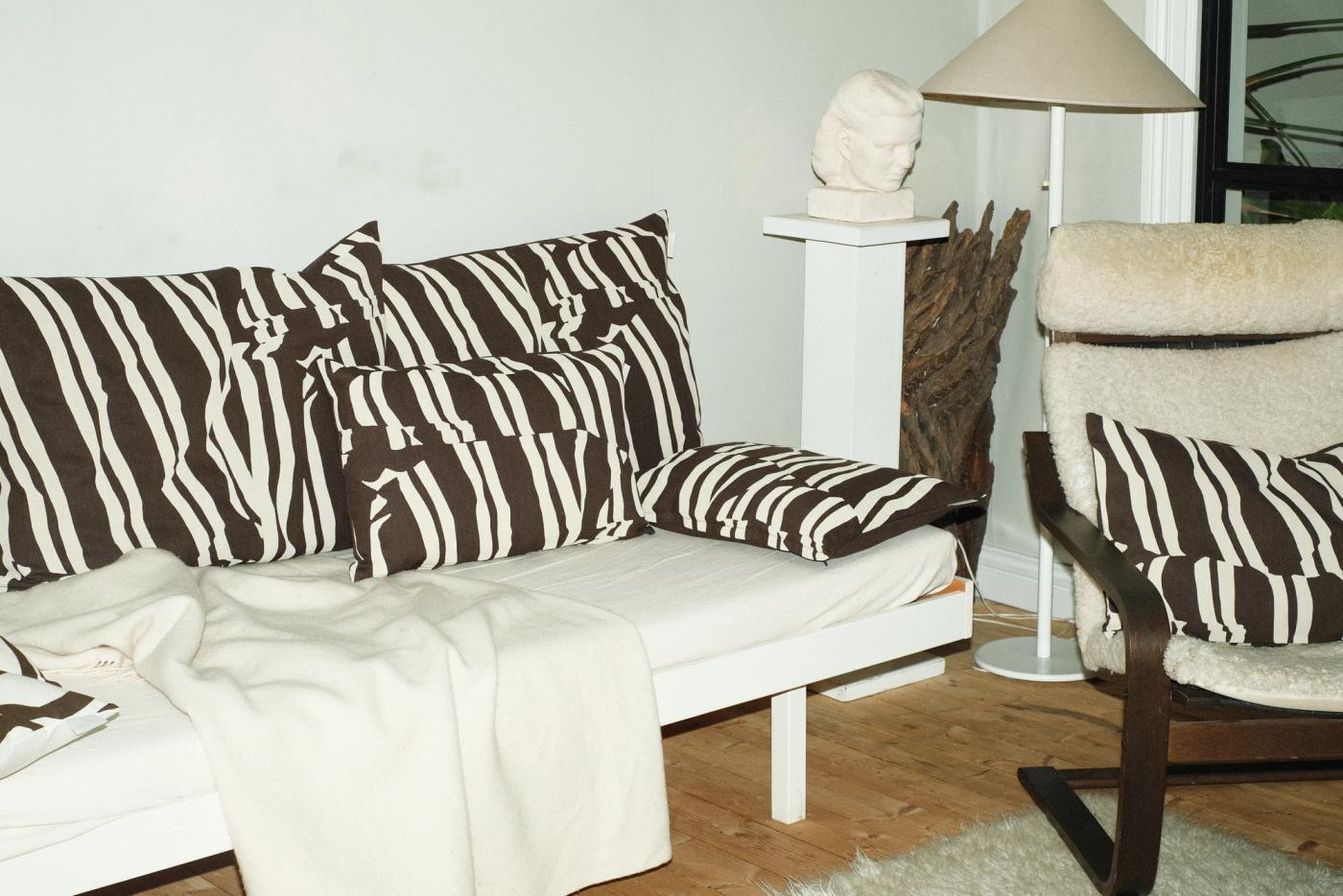 A light-toned sofa and a sheepskin armchair with several black-and-white striped cushions, with a white sculpture and houseplant in the background.