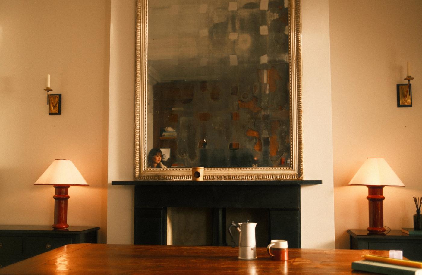 Two red table lamps lit on both sides of the fireplace, with a large mirror above it and a wooden table in front of it.