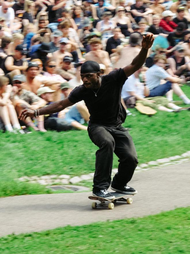 A skateboarder crouches low with arms outstretched for balance, watched by a big crowd sitting on the grass