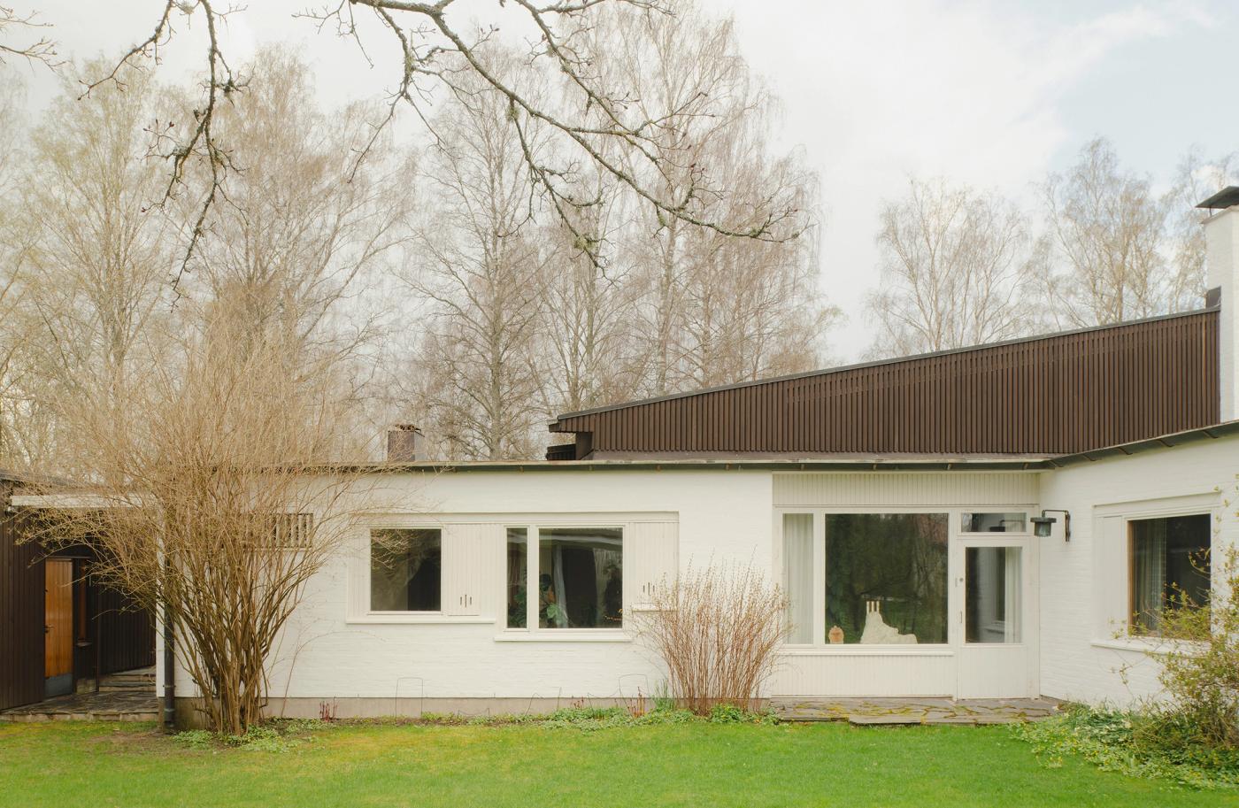 Villa Skeppet seen from the backyard, showing the L-shaped floor plan, white rendered facade, dark sloping roof, and surrounding birch trees