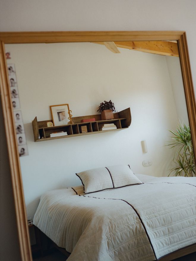 A view reflected in a large mirror into the bedroom: a white bed, a wooden wall shelf, and a houseplant.