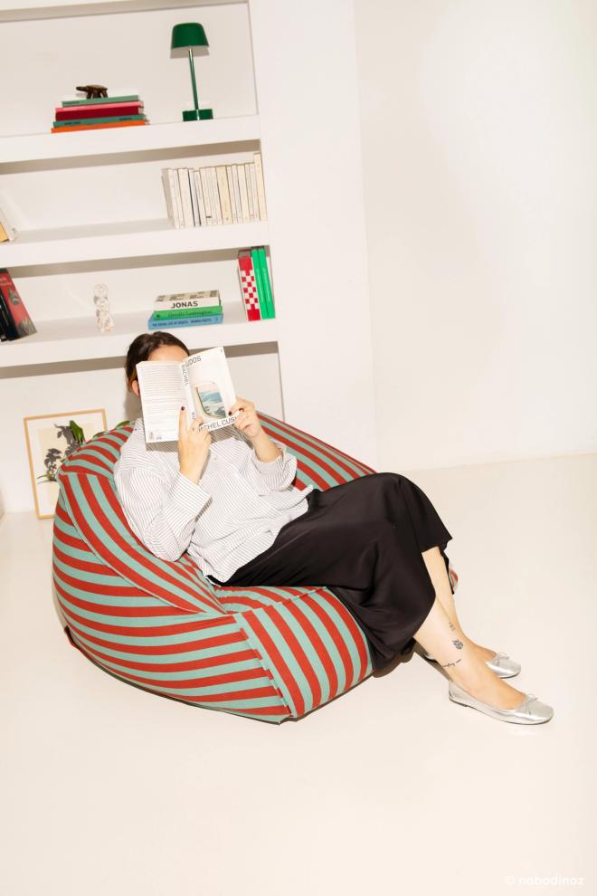 A person reading a book, half-reclined in a red-and-white striped beanbag in front of a white bookshelf