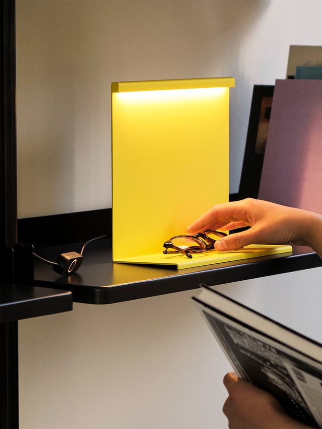 LBM table lamp in yellow with a hand reaching from the right to place eyeglasses on top of the horizontal metal sheet at the bottom of the lamp.
