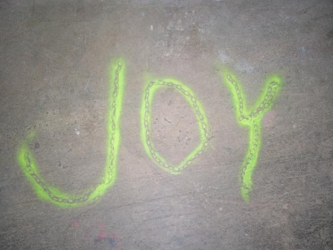 The word 'JOY' spray-painted in neon green on a concrete floor
