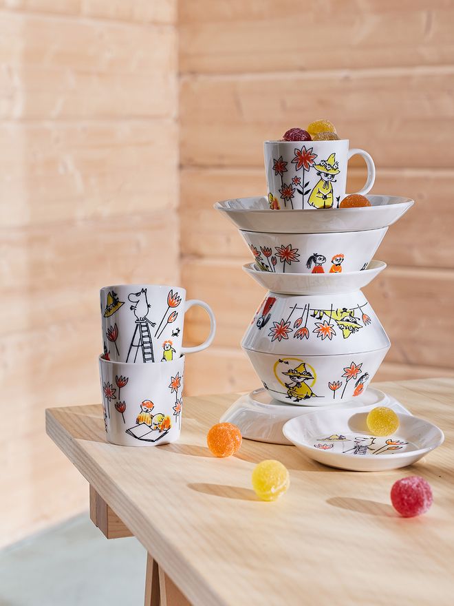 A quirky pile of Moomin by Arabia x Red Cross tableware on a wooden table: mugs, plates, bowls.