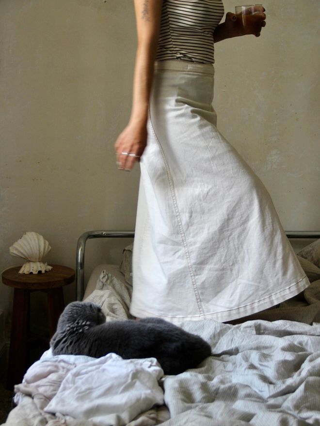A woman in a long white skirt stands on a a chrome-framed bed, a black cat curled up on the bedding in front of her.