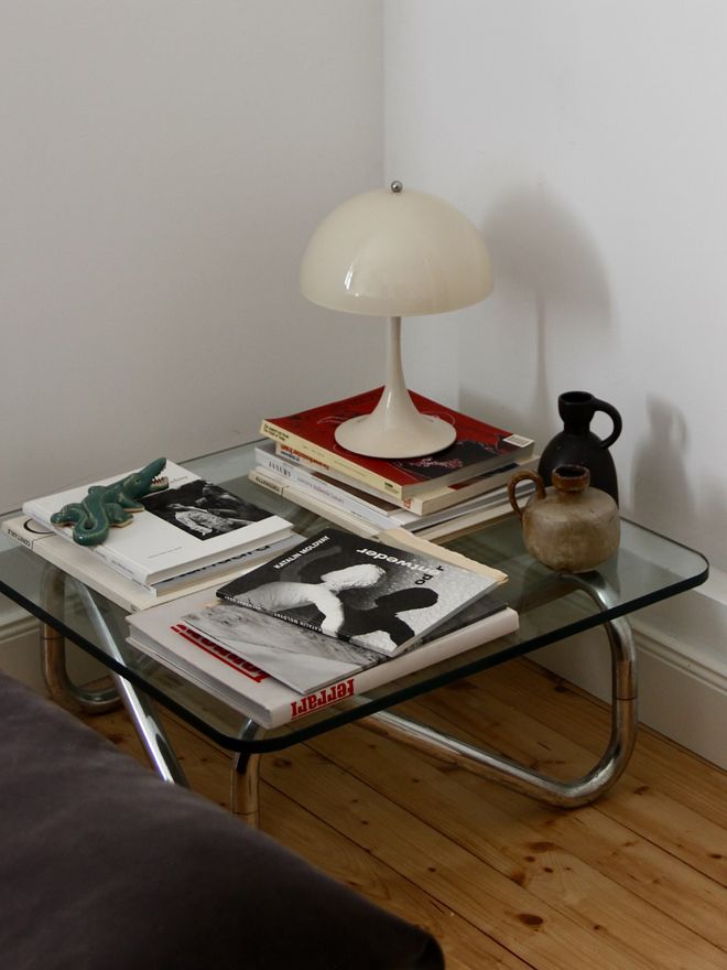 A glass coffee table with a pale Panthella table lamp, a stack of books, and a dark stoneware jug.