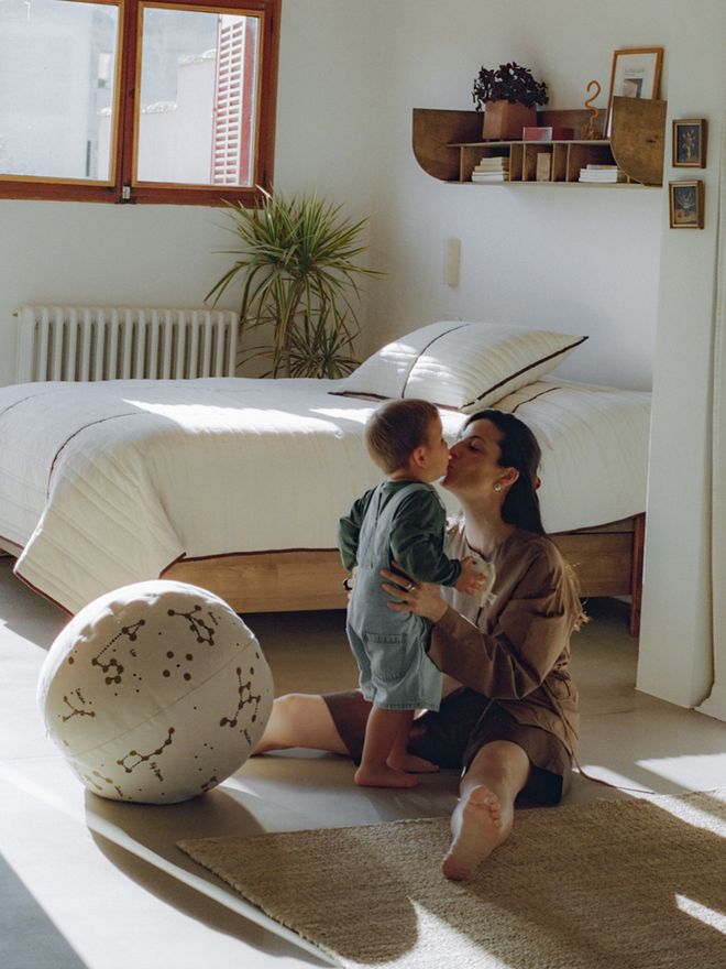 Cecilia sits on the bedroom floor, kissing a child on the cheek. A wooden bed with pale linens and a large houseplant stands behind them.