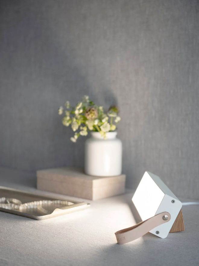 White Valovoima Mini light therapy lamp on a light-colored table, with a vase of small white flowers in the background.