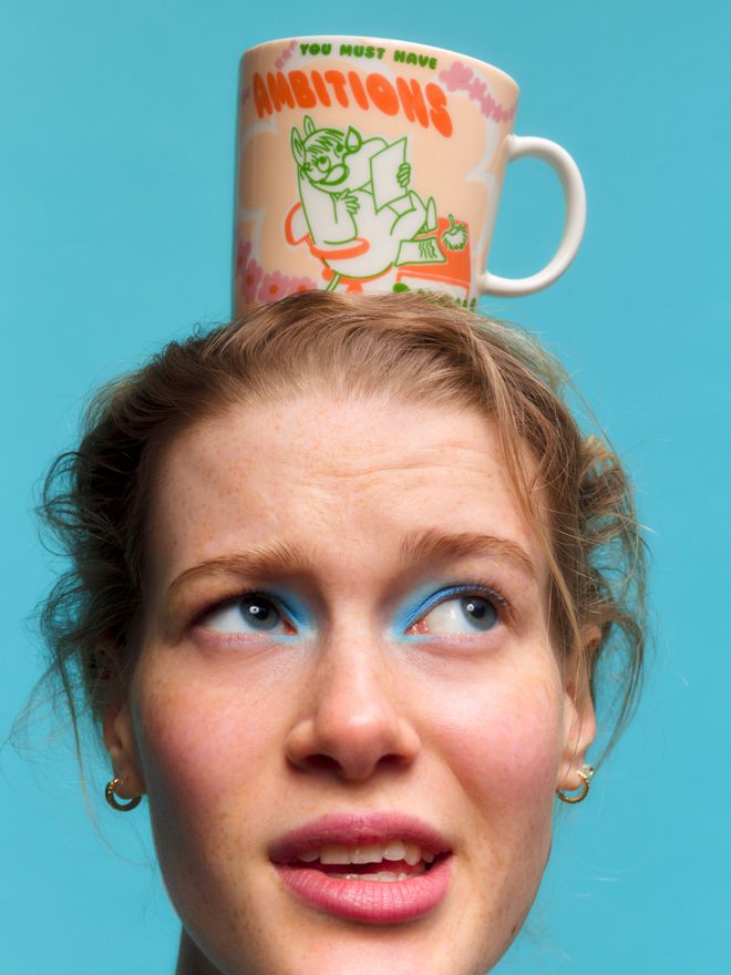 A person is balancing a patterned “Boss Lady” Moomin mug on their head against a blue background, looking up.