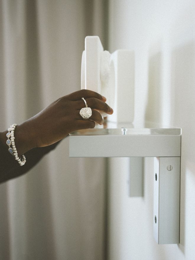A hand wearing a diamond ring and pearl bracelet touches white objects on a wall shelf