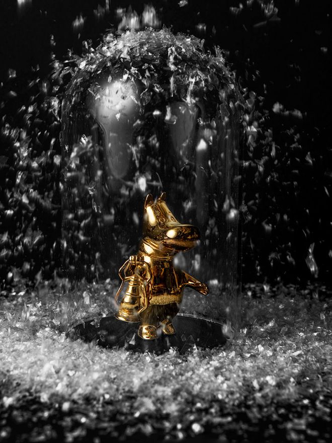 A gilded Moomintroll figurine carries a lantern in a snowfall.