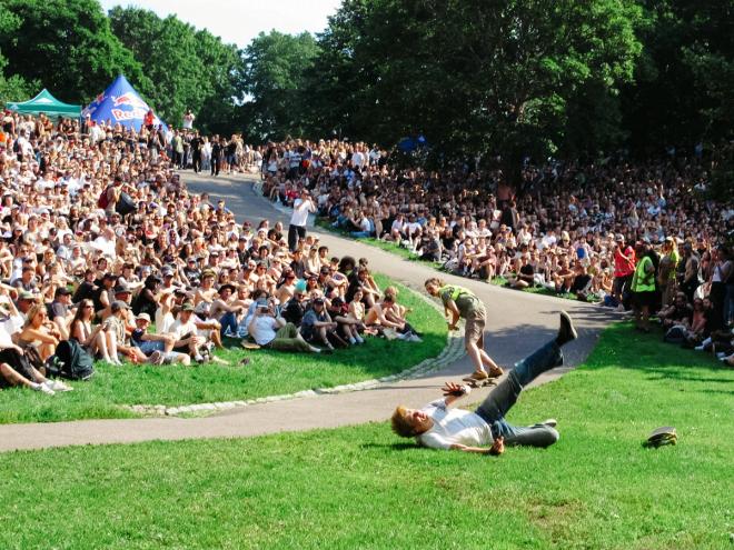 A skateboarder lies fallen on his back on the grass at the end of the track, board flung aside, as another rider comes down behind and a large crowd watches from the slope.