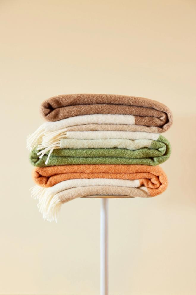 A stack of folded fringed wool blankets in brown, beige, olive green, orange, and natural white