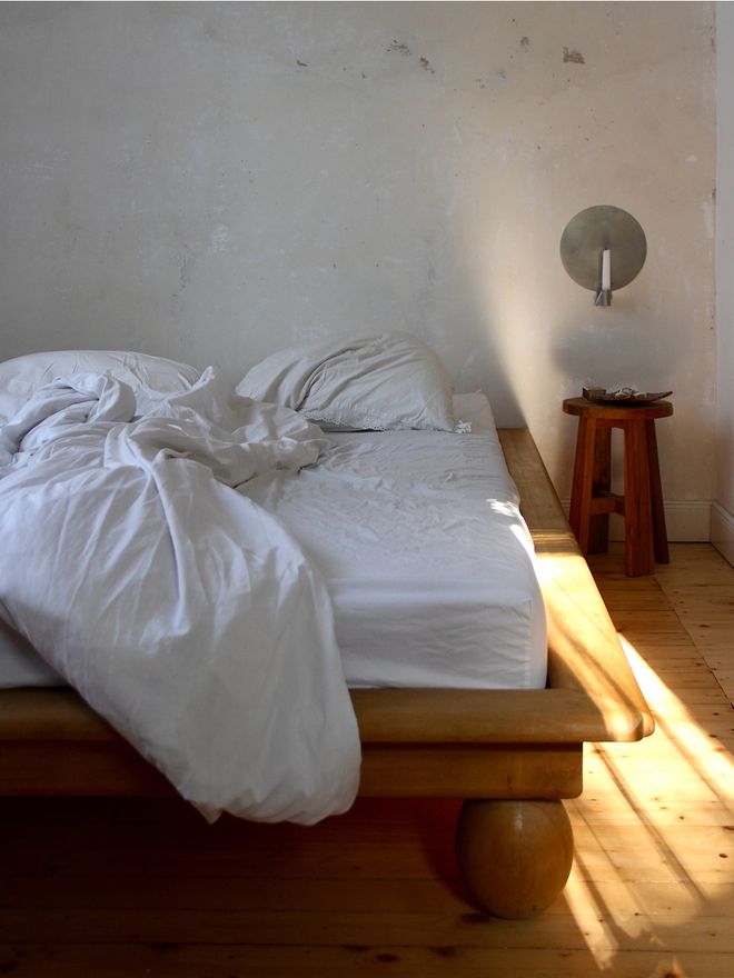 Rumpled white bedding on a low wooden bed, a rough white wall with a small wall candle holder, a wooden stool, and sun on the plank floor."
