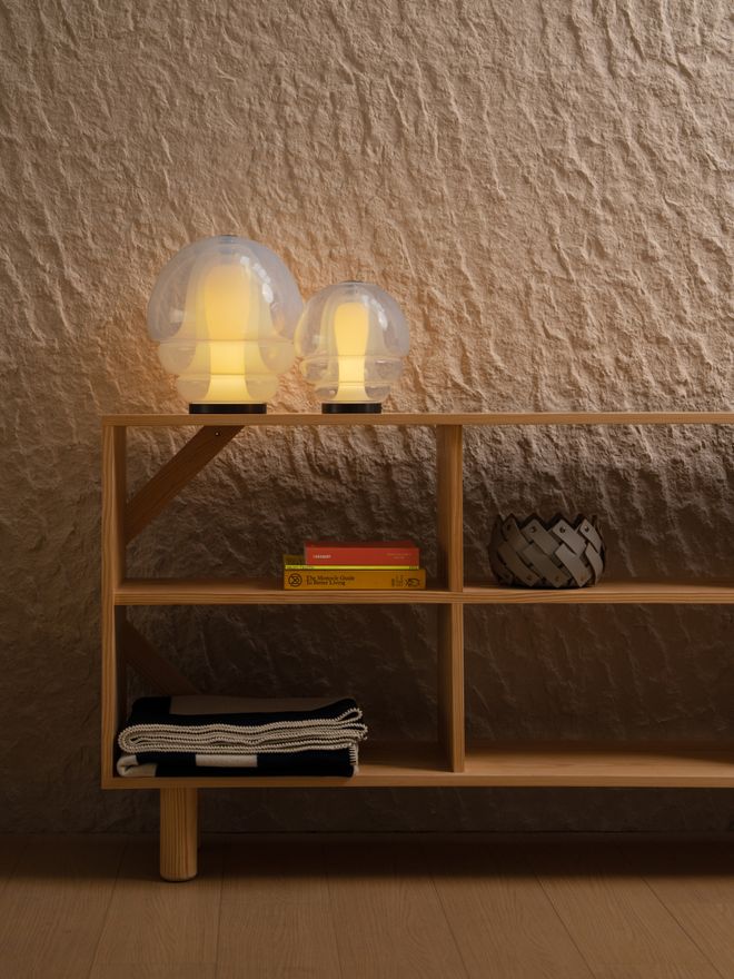 Two lit Medusa table lamps on top of a wood shelf against a textured plaster wall. The shelves below hold a stack of books, a decorative bowl, and a folded black-and-white textile.