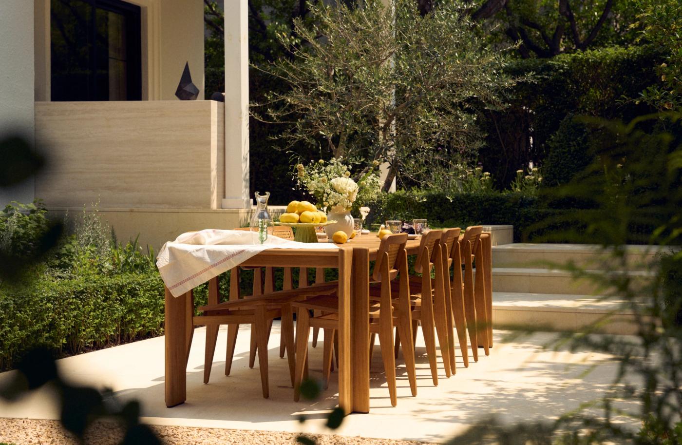 Long wooden dining table with chairs on a sunlit stone terrace, set with lemons and white flowers, with an olive tree and hedges behind it.