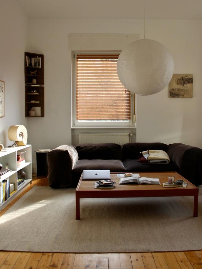 A light living room with a dark sofa, a low wooden coffee table, a bookshelf, and a large round paper lantern overhead.