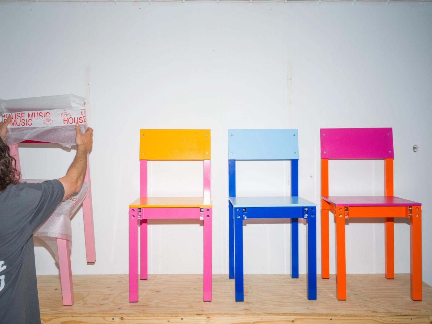 Three two-tone chairs stand side by side on a wood floor — pink-and-yellow, blue-and-light-blue, and orange-and-magenta; at left, Fredrik Paulsen himself covers a fourth, pink chair with a cloth printed 'House Music'