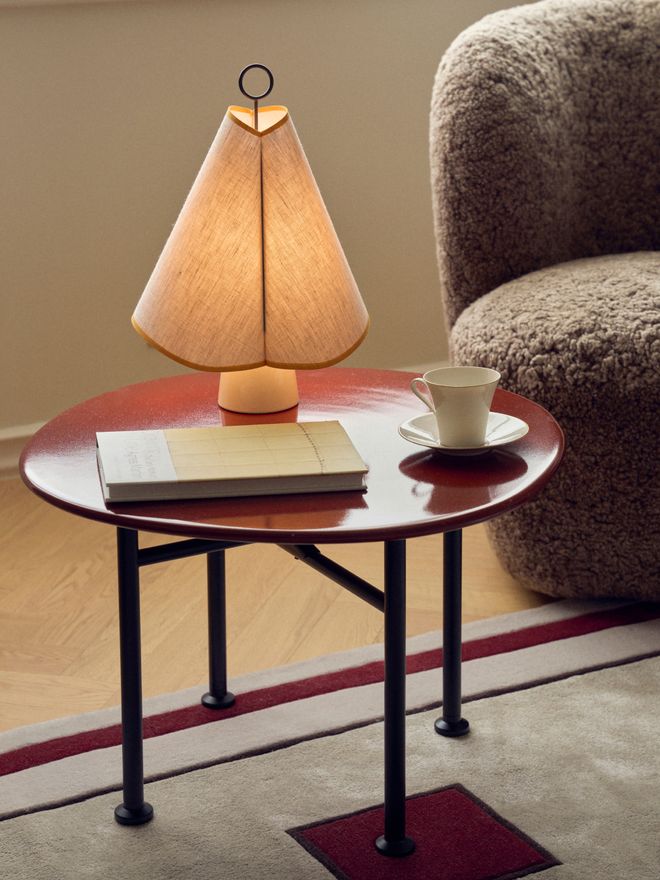 Close-up of a lit table lamp with a pleated fabric shade and a ring handle on a round red side table, next to a book and a teacup.