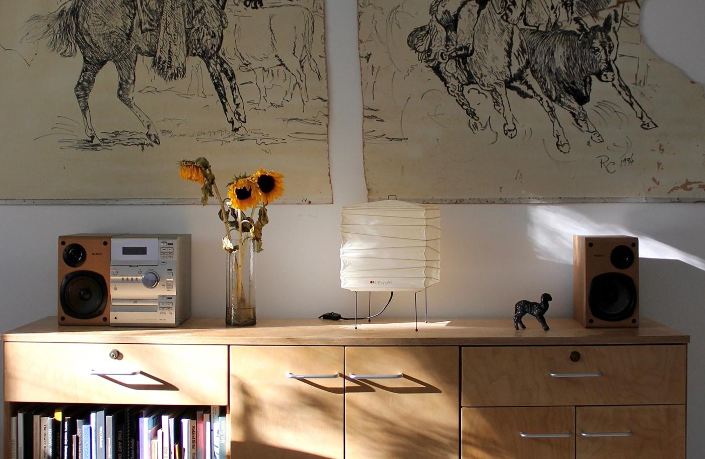 A wooden sideboard with a stereo, speakers, and a bouquet of sunflowers, above hangs a large drawing of horses and riders.