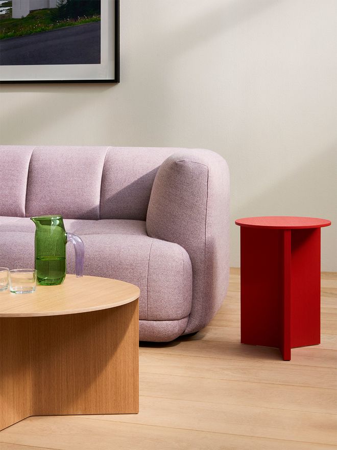 A red Slit Wood table next to a lilac sofa, and a wood-colored Slit Wood table in front of the sofa. On the table are a green water jug ​​and two drinking glasses.