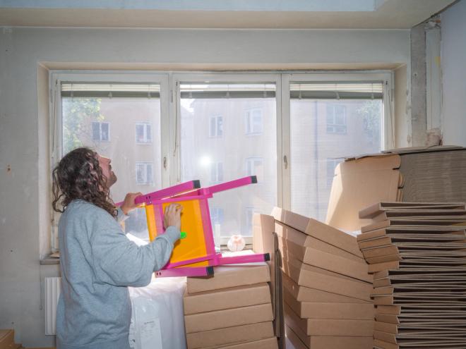 Fredrik Paulsen is examining a chair with a pink frame and yellow seat in his workshop, surrounded by stacks of flat-packed cardboard boxes.