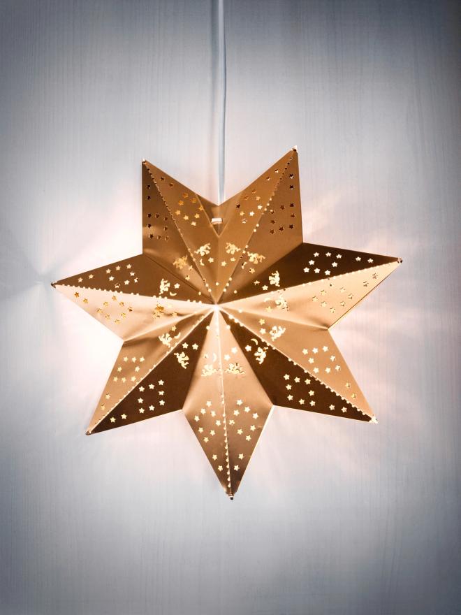 A lit gold metal star pendant hanging against a wall, its perforations showing as points of light.