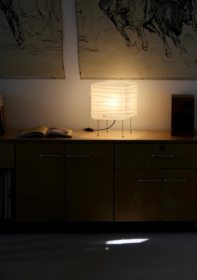 A lit Akari paper lamp on a wooden cabinet in a darkened room, with a horse drawing hanging on the wall above.