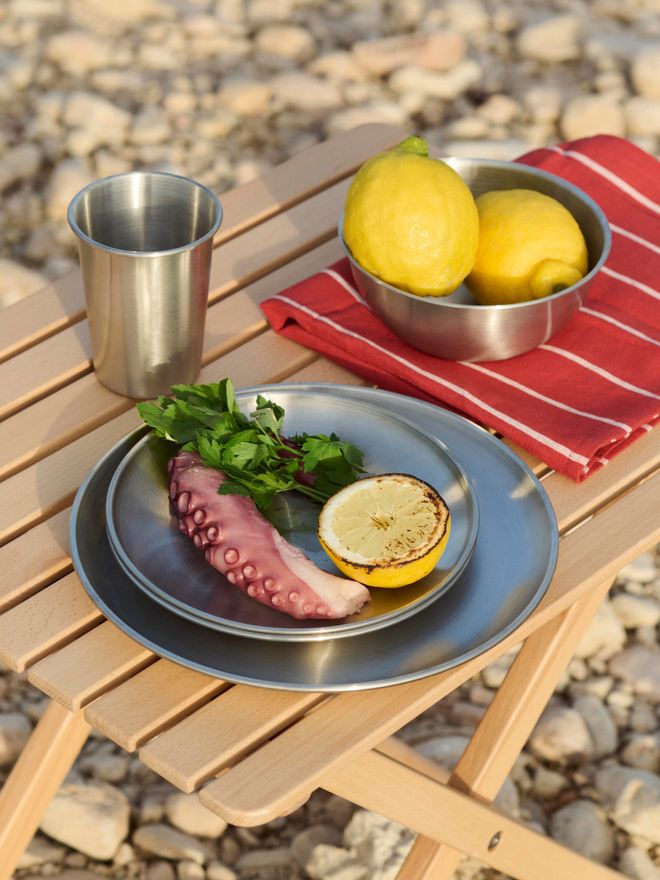 A metal plate on a folding table, topped with a squid tentacle, parsley, and half a lemon, with a steel mug and a bowl of lemons placed next to it on a red cloth.