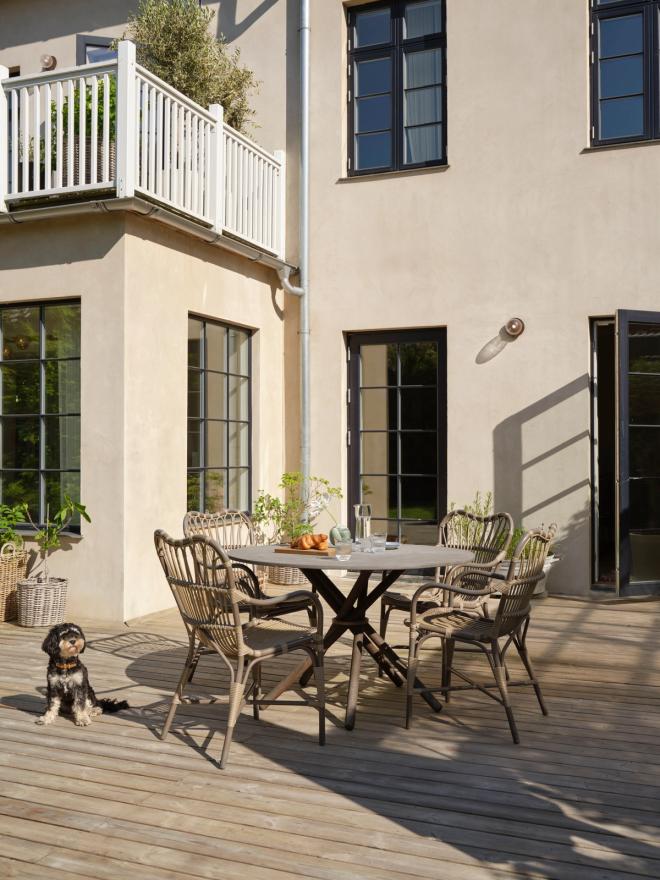 A wooden terrace with a round dining table and rattan chairs behind the villa, and a small black dog resting on the deck.