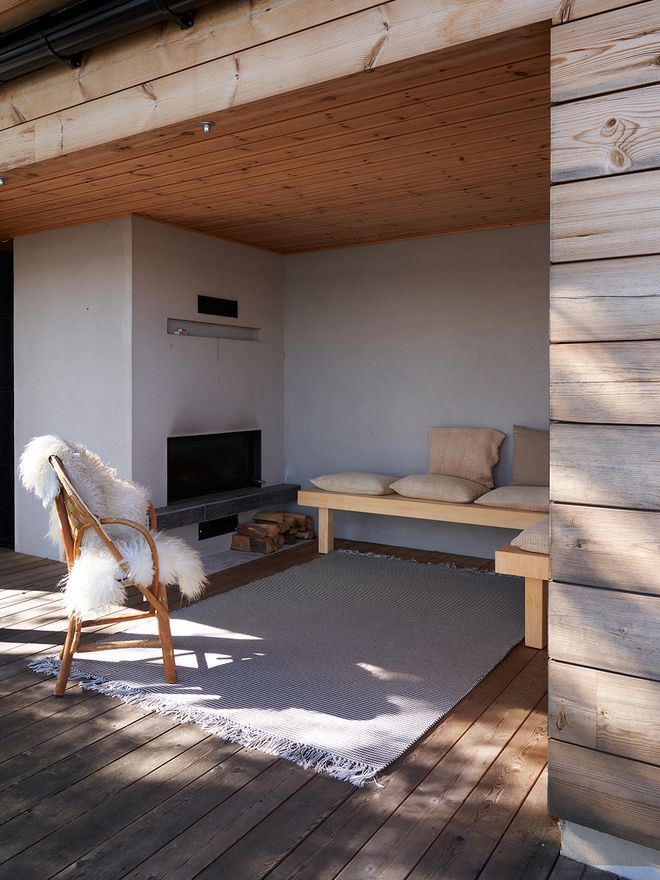 An outdoor fireplace area with built-in benches next to the lower-level sauna, sheltered by the upper terrace above. A Woodnotes Grain In-Out rug suitable for outdoor use on the floor.