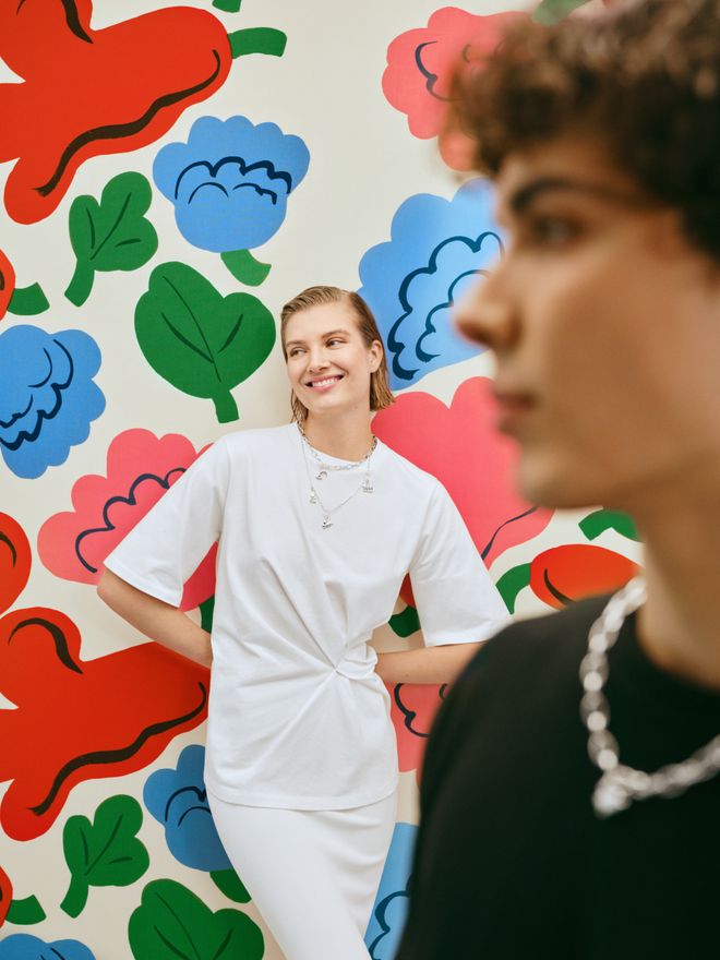 Two people in front of a colourful floral wall, one in black in the foreground and one in white smiling in the background