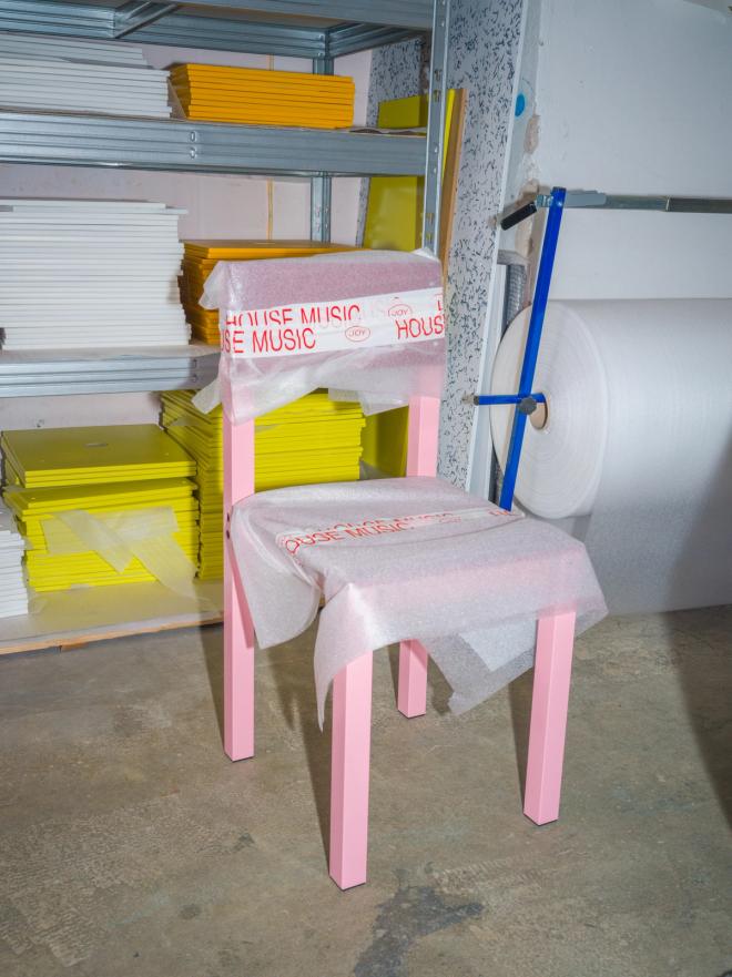 A pink chair stands wrapped in paper printed with red 'House Music' text, in a workshop with shelves of yellow and white sheet material and a roll of plastic film.