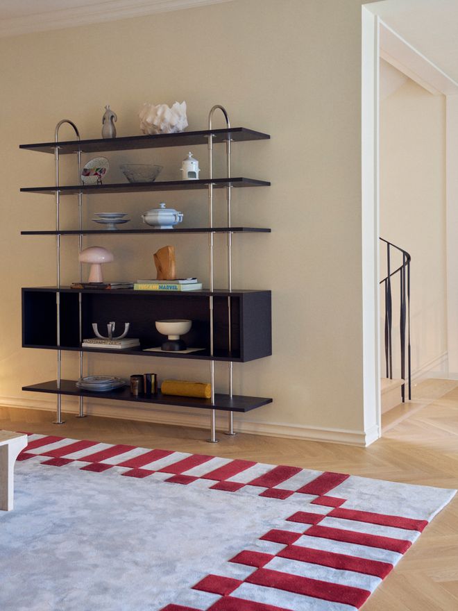Dark shelving unit holding ceramics, bowls, and vases, against a beige wall beside an open doorway, with a red and white patterned rug on a herringbone wood floor.