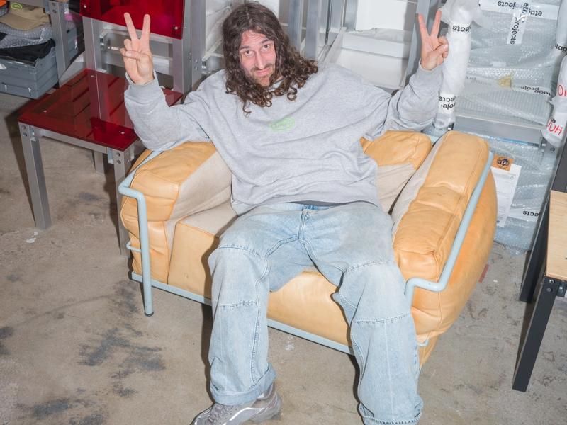 Fredrik Paulsen flashes a double peace sign while lounging in a beige armchair in his colorful studio.