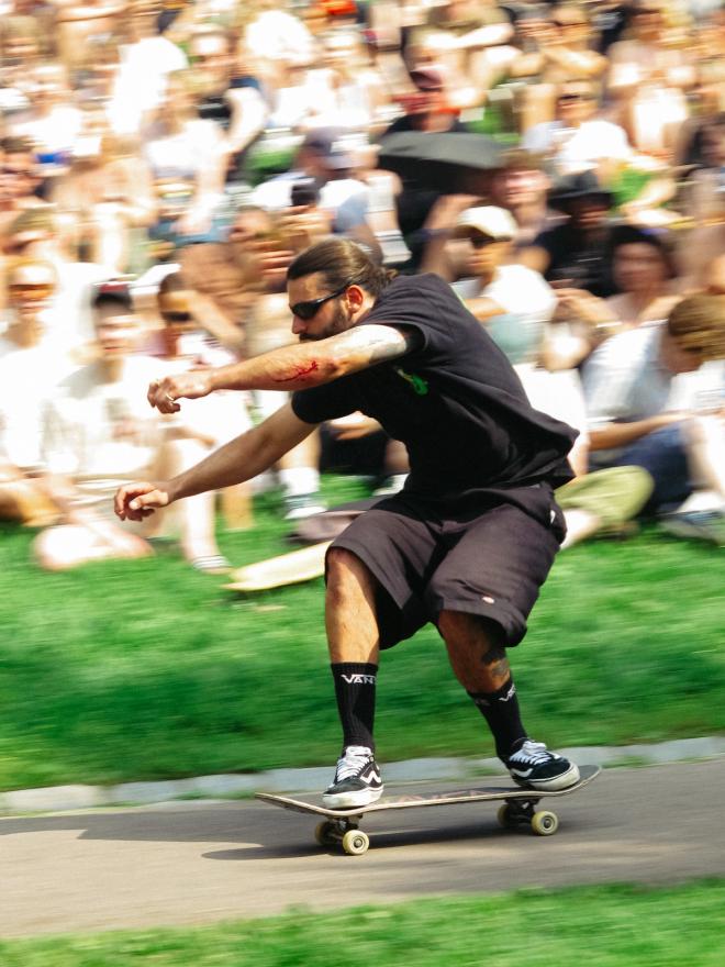 A bearded skateboarder crouches low on his board, racing past a blurred crowd