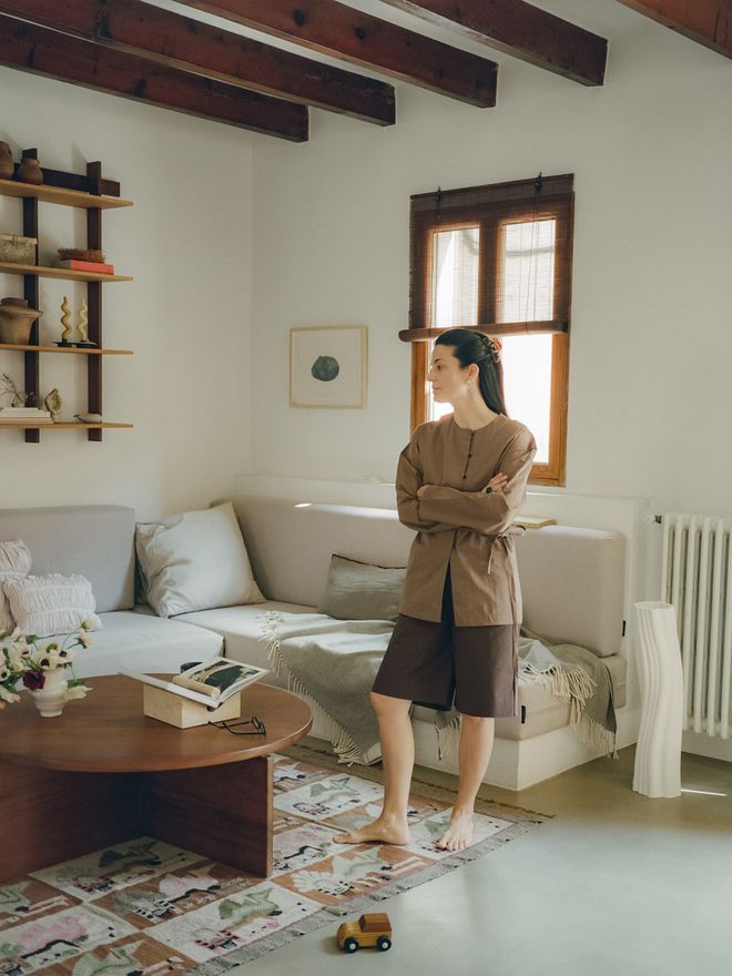 Cecilia stands in a light-filled living room with wooden ceiling beams, a pale corner sofa, and a round coffee table behind her.
