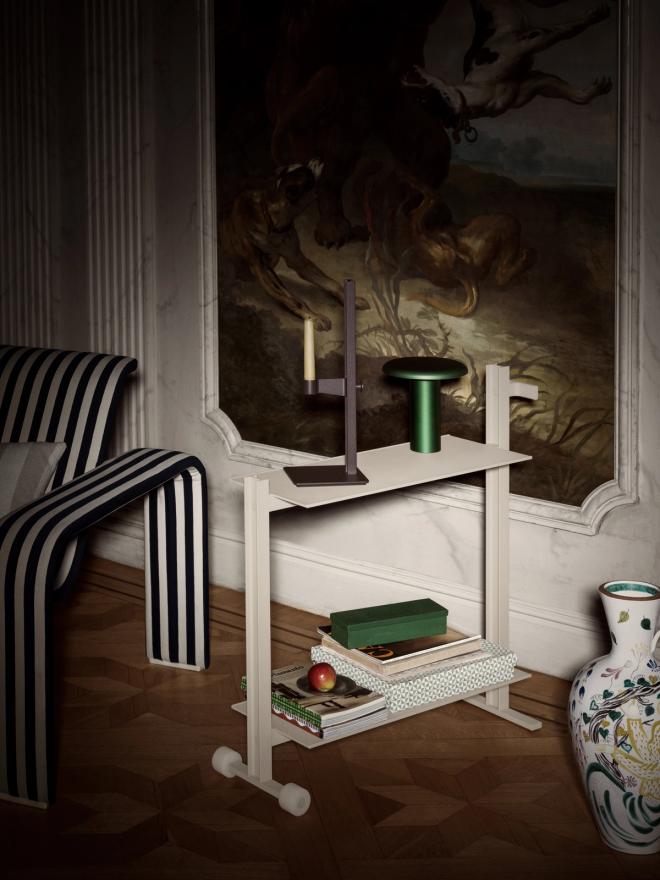 A white two-tier trolley with a candleholder and a green table lamp on the top shelf and books on the lower shelf