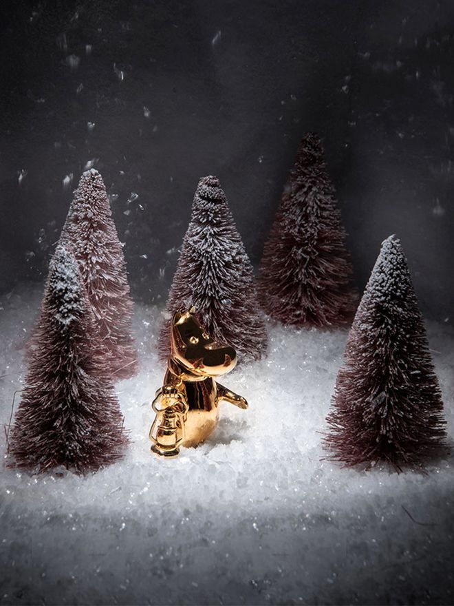 A gilded Moomintroll figurine carries a lantern in a snowy landscape.