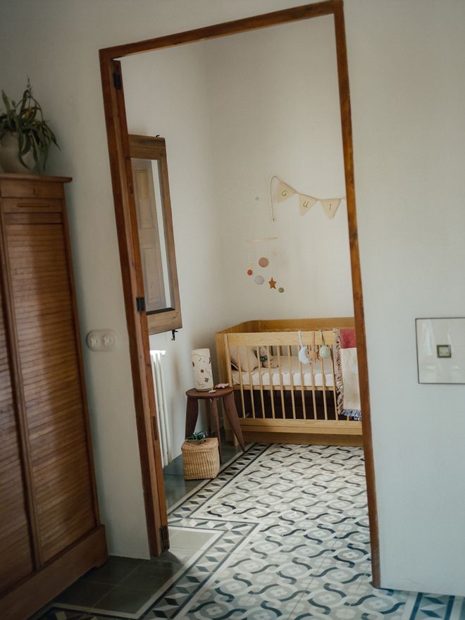 A view through a doorway into the nursery: a wooden crib, a pennant garland on the wall, and the original patterned tile floor.