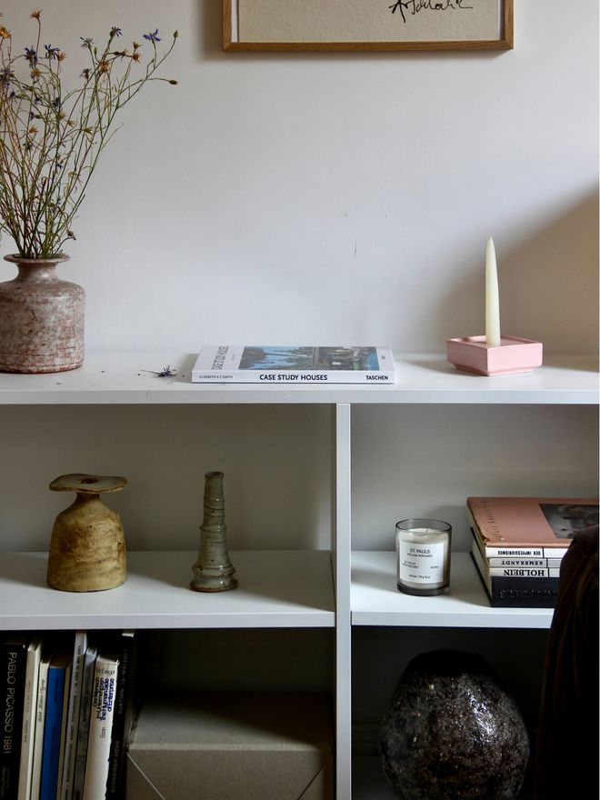 A white bookshelf with books, ceramic pieces, candles, and a bouquet of dried flowers.