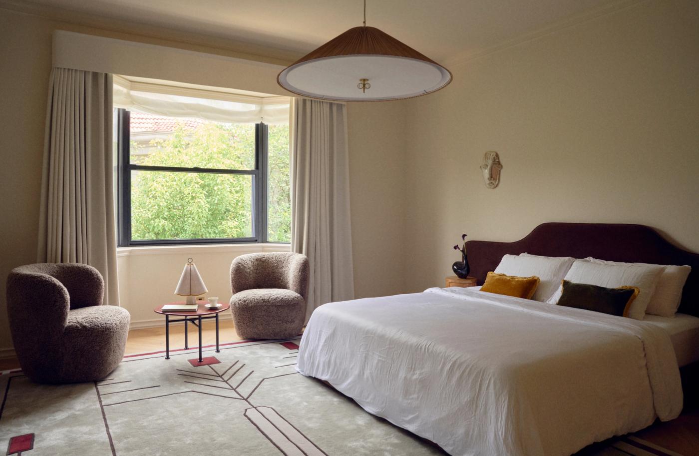 Bedroom with a white bed and dark headboard, two textured armchairs and a small table by a window overlooking green trees, and a patterned rug on the floor.