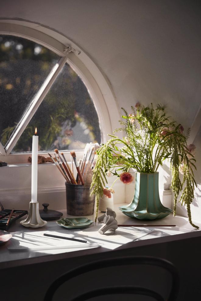 A still life arranged in front of an arched window, featuring a burning candle, brushes in a cup, a vase of flowers, and drawing supplies