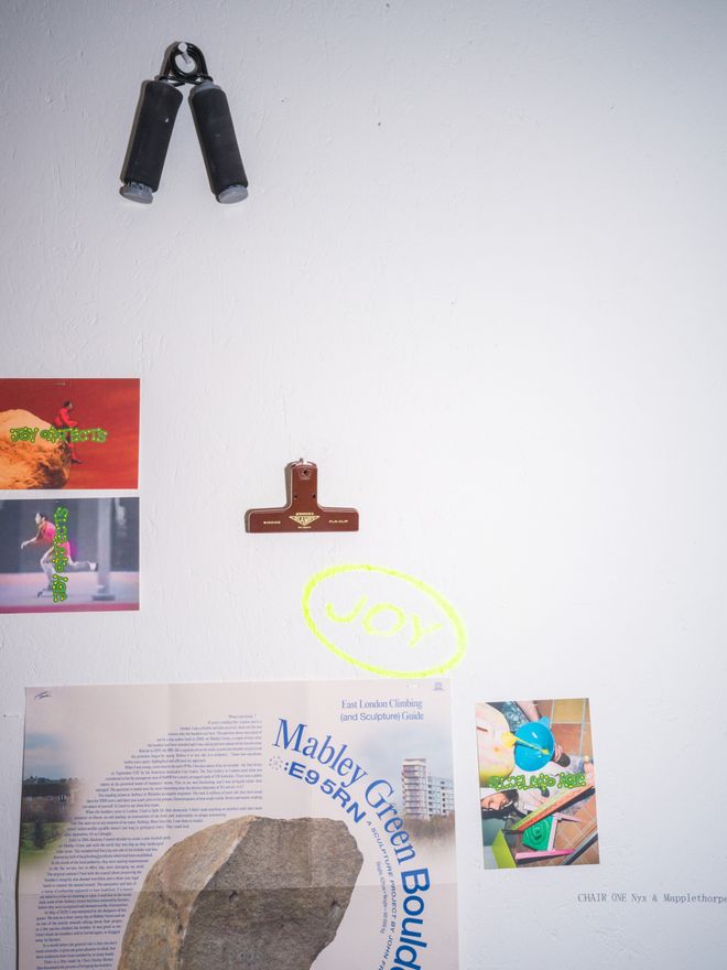 A cluttered wall collage of photos, clippings, and small objects, including a hand grip trainer and a wooden ornament