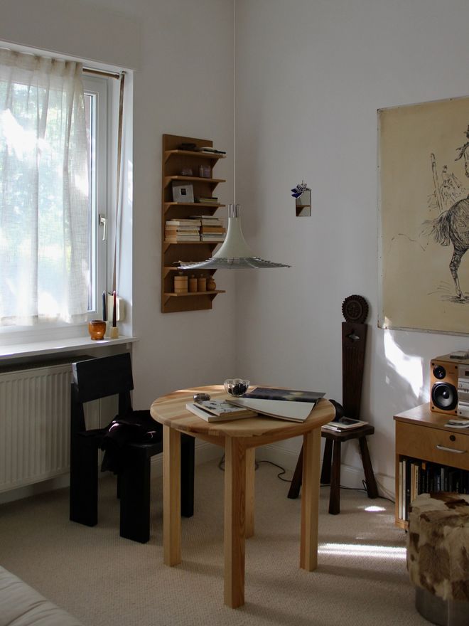 A light-filled room with a black Vaarnii wooden chair at a round wooden table, and a window with curtains and a small wall shelf in the background