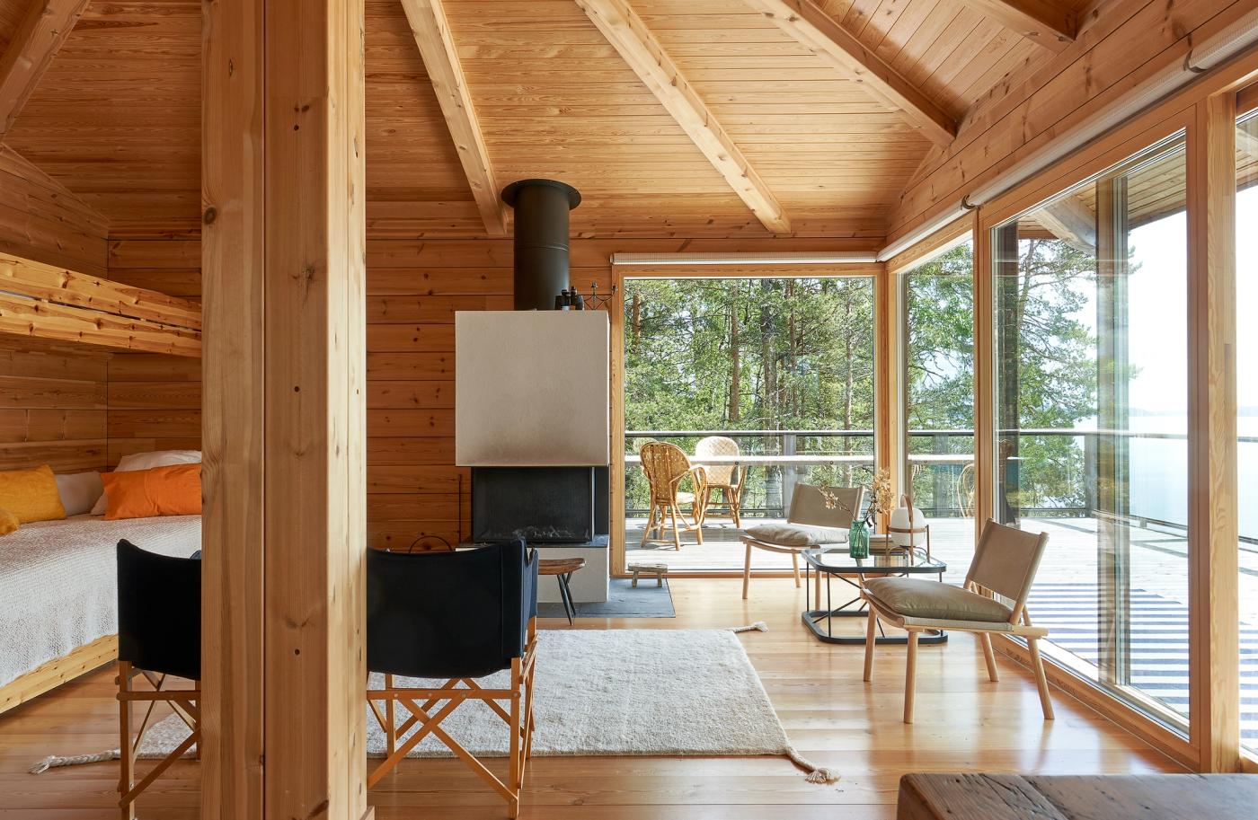 The bright living room of the cabin, with the exposed hip roof structure, a continuous row of windows facing the lake, and a Woodnotes Uni wool rug on the floor.